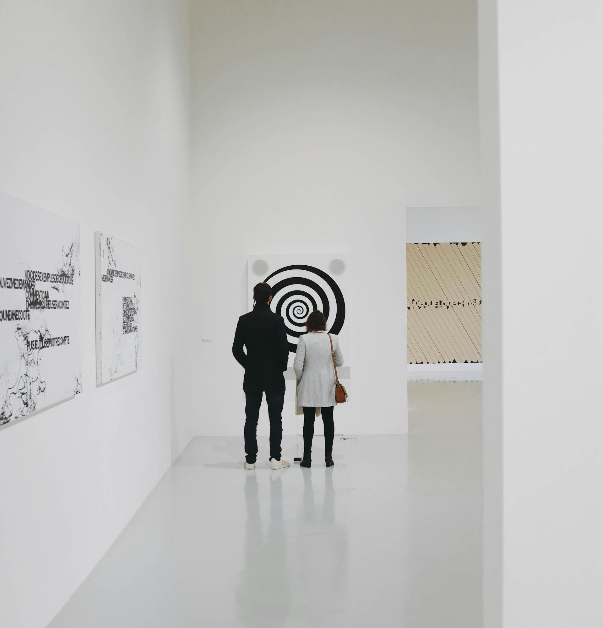 Two people standing in a bright, minimalist art gallery observing a black-and-white spiral artwork, with additional contemporary pieces displayed on surrounding white walls.