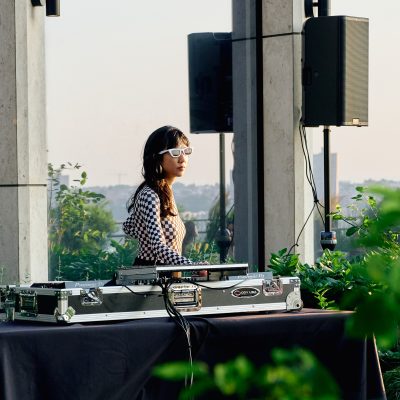 A DJ with white sunglasses and a checkered shirt performs at a rooftop turntable setup surrounded by greenery.