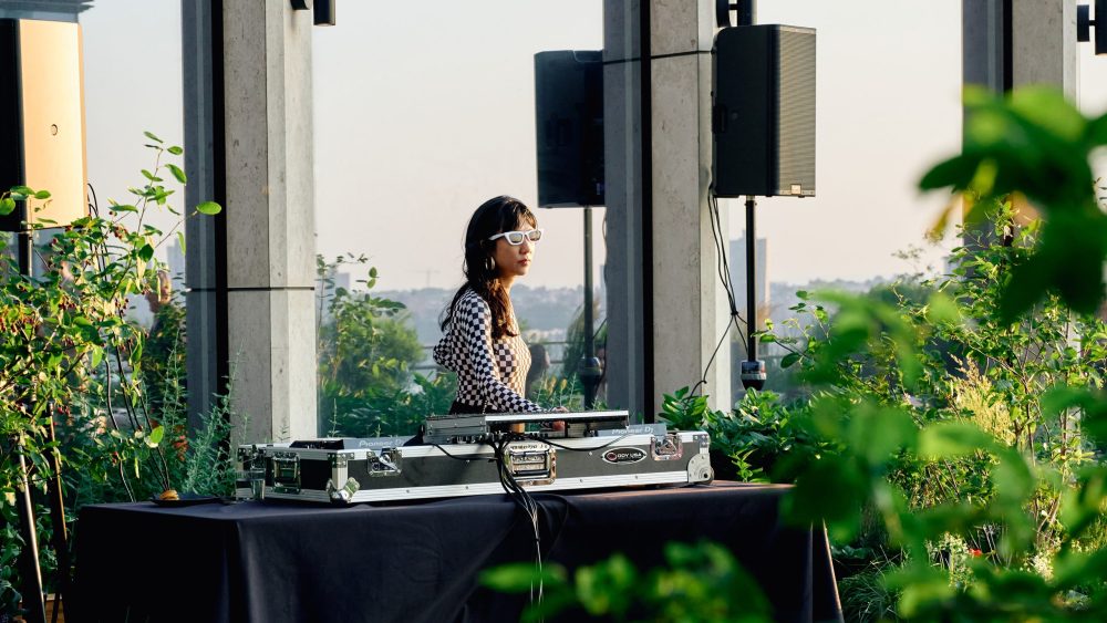 A DJ with white sunglasses and a checkered shirt performs at a rooftop turntable setup surrounded by greenery.