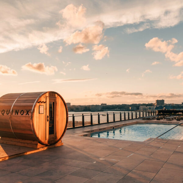Rooftop pool at sunset with lounge chairs, barrel saunas, and city views for a serene wellness escape.