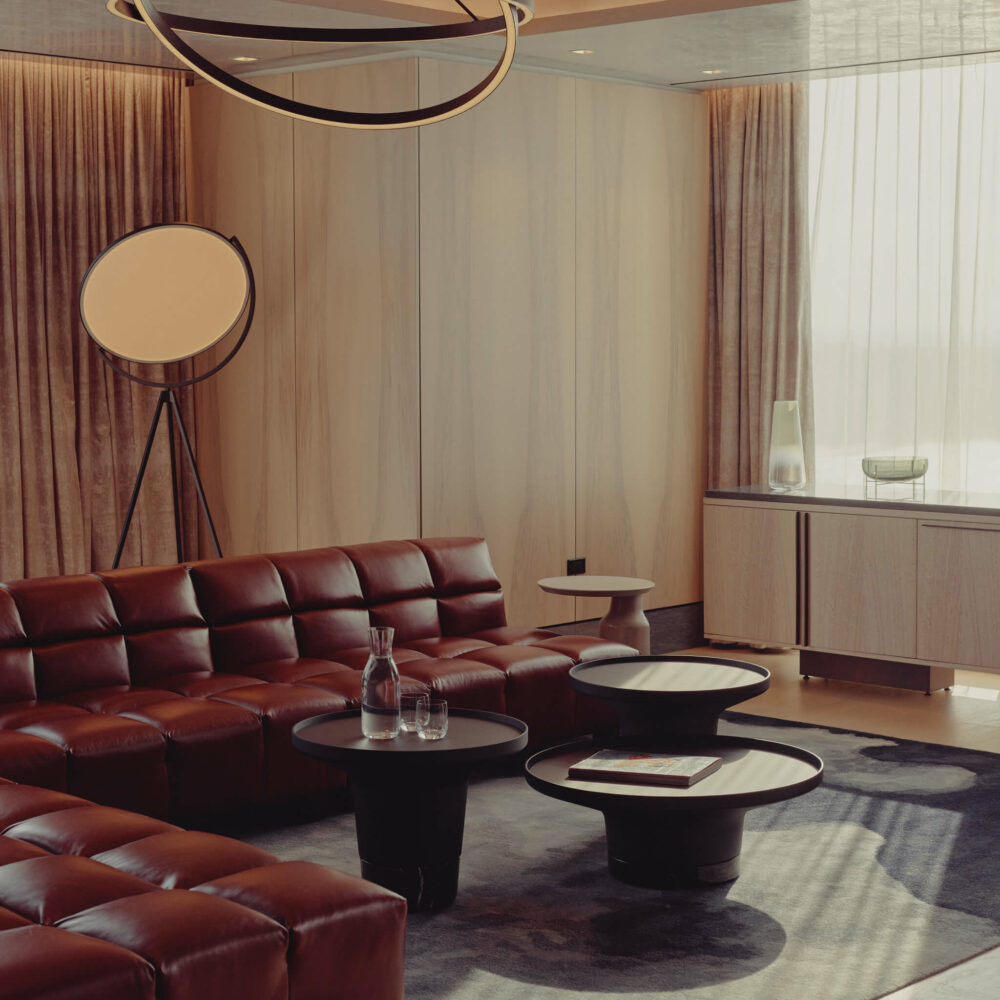 Modern living room with dark leather sectional sofa, round coffee tables, wood paneling, and geometric chandelier.