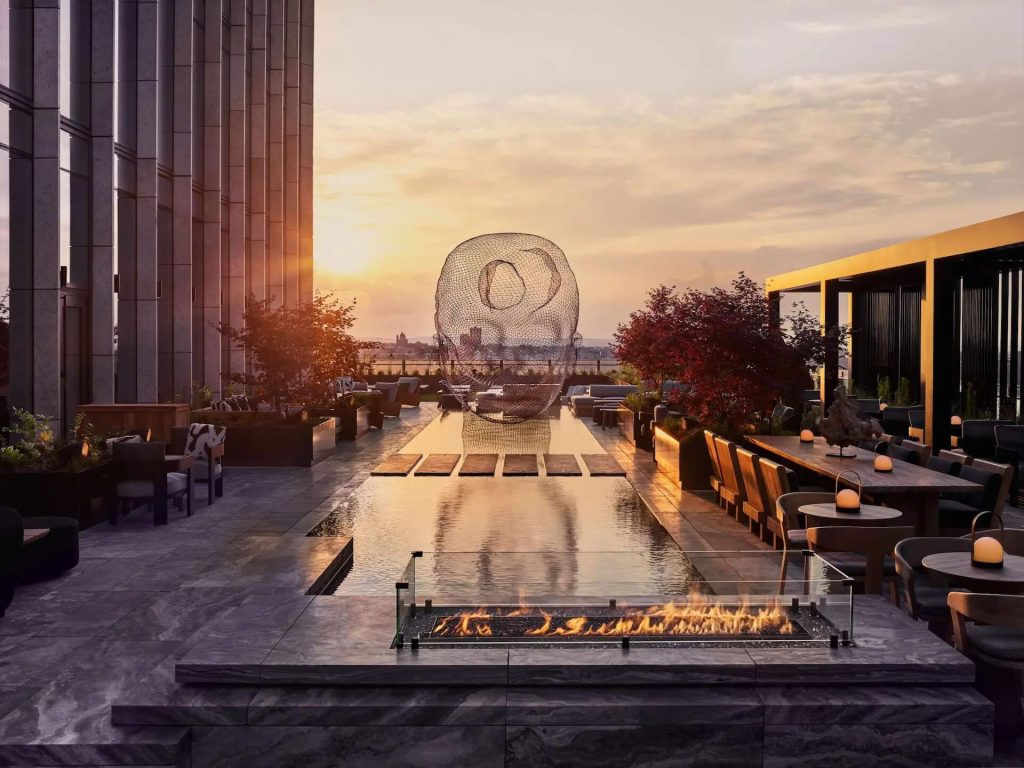 Rooftop terrace at sunset with reflecting pool, wireframe head sculpture, and warm seating area.
