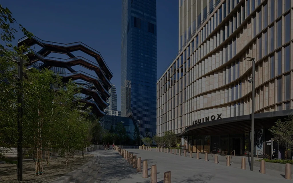 View of the Vessel and Equinox Hotel in NYC, framed by trees and modern skyscrapers in Hudson Yards.