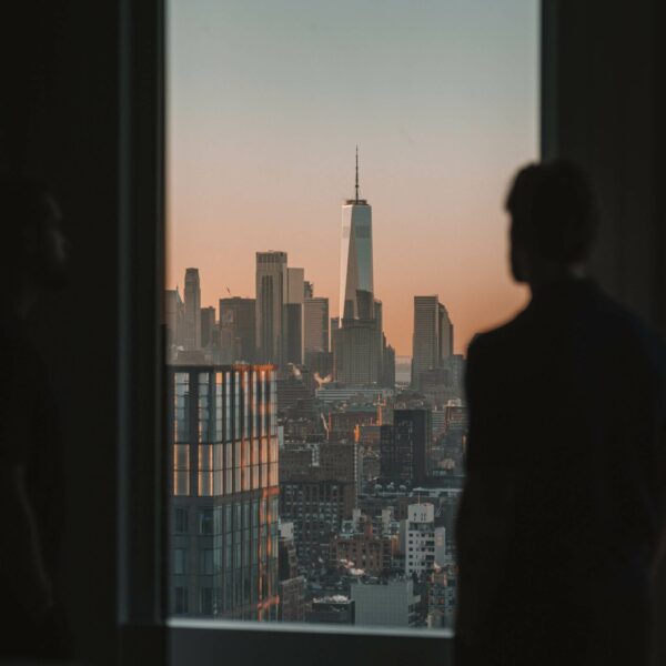 A man stands by a window at sunset, gazing at New York’s glowing skyline and tall luxury buildings.