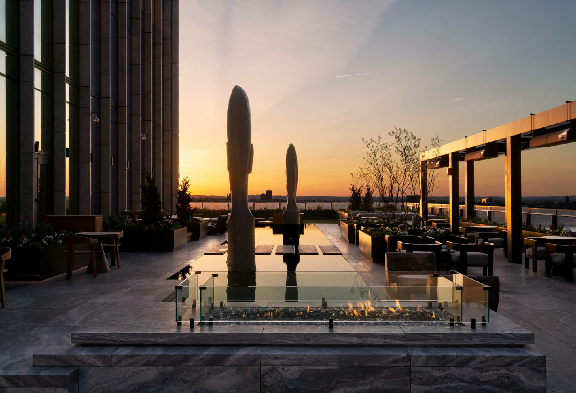 Rooftop terrace at sunset with abstract sculptures, water feature, and city views in a serene setting.