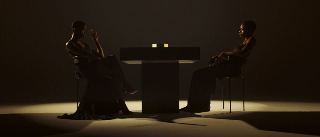 Two people sit across a table in a dark room, lit by a glowing cube that creates a tense, minimalist mood.