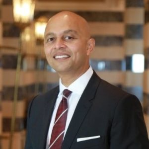 Man in dark suit with red striped tie standing indoors against elegant striped wall background.