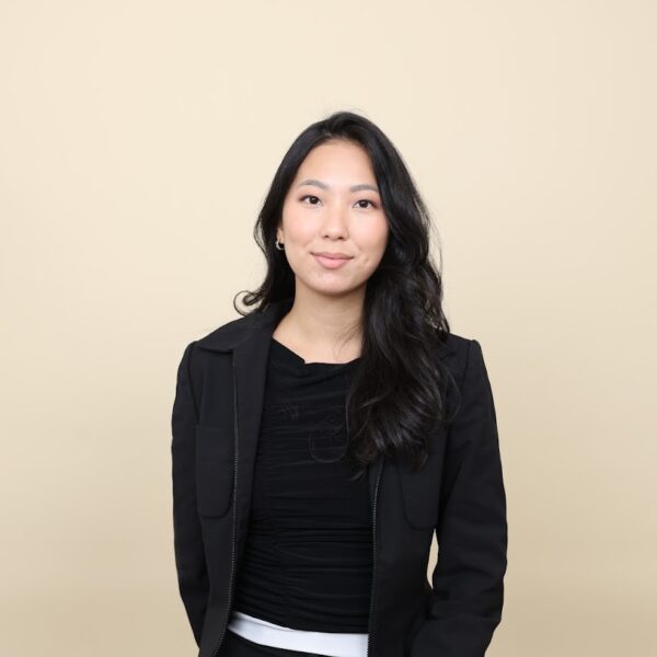 Tara Laksmono, seated in black attire against beige backdrop, exuding poise and professional elegance.