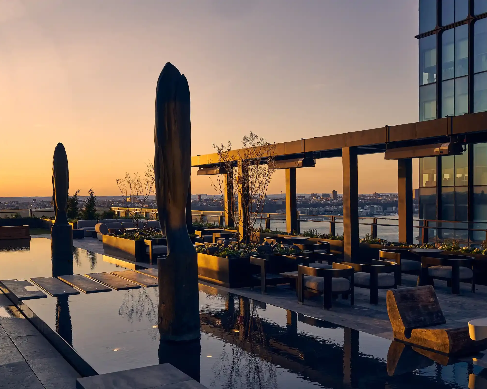Rooftop terrace at sunset with reflecting pool, sculptural pillars, and pergola seating overlooking city.