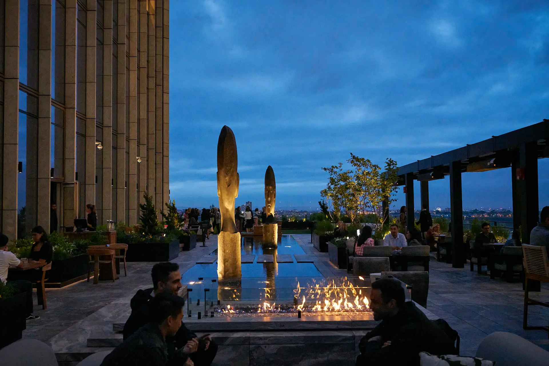 Elegant rooftop lounge with sculptures, fire pit, and city views, perfect for dining and connection.