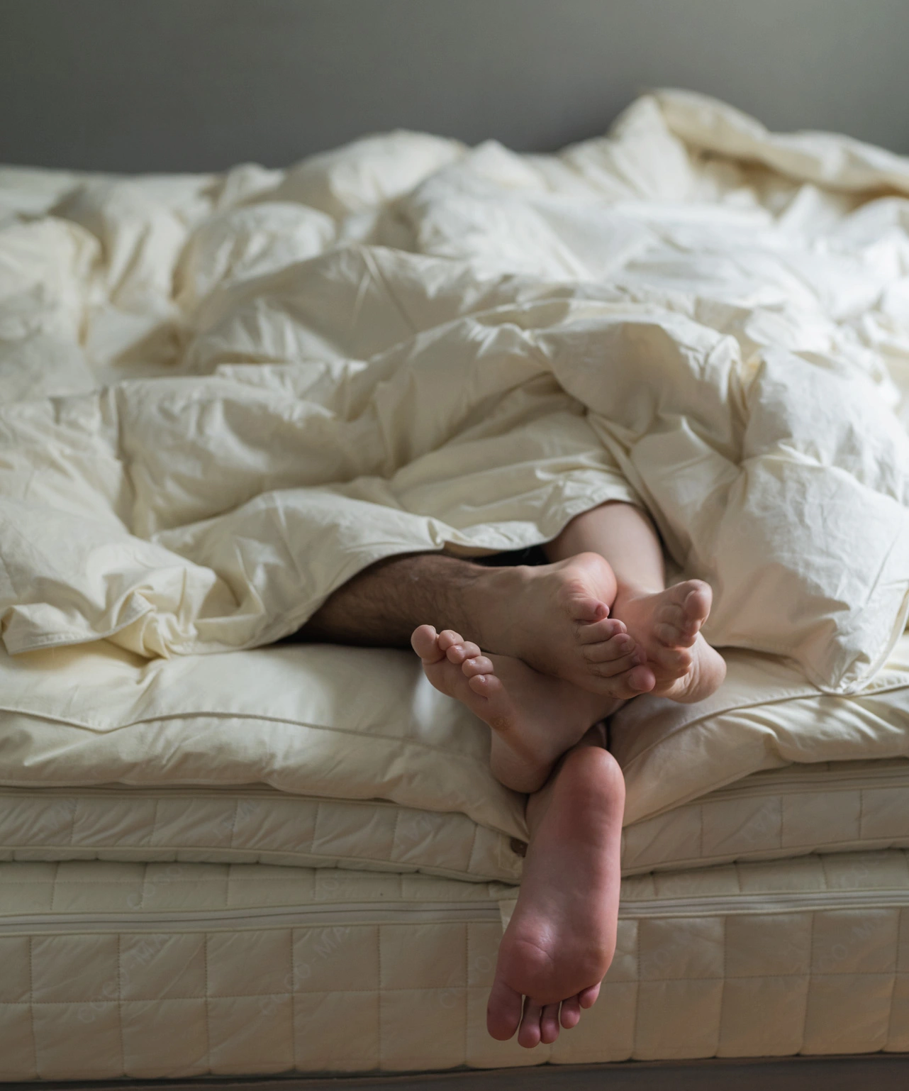 Two guests resting together under cream-colored bedding, feet peeking out in cozy, softly lit bedroom