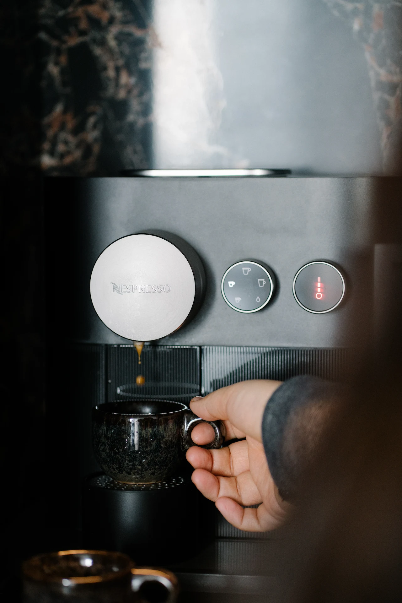 Guest brewing espresso with Nespresso machine on dark marble counter, holding glass cup beneath spout