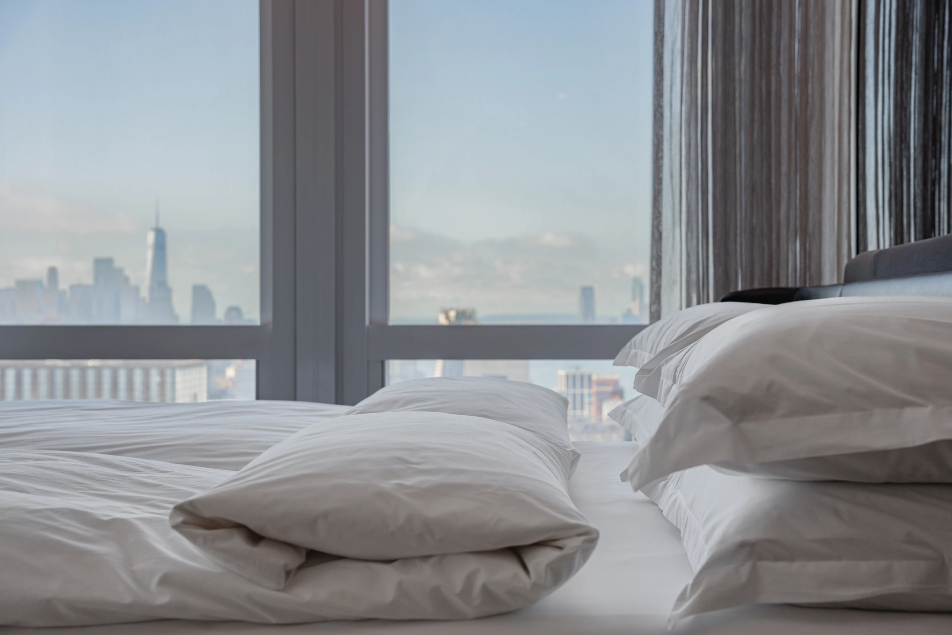 Minimalist bedroom with white bedding and panoramic city skyline view through floor-to-ceiling windows, bathed in natural light