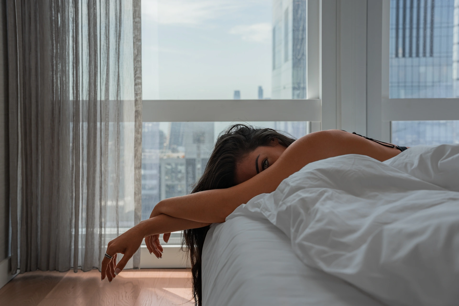 Woman lies on bed near window, partly covered, one eye visible, daylight, cityscape behind.