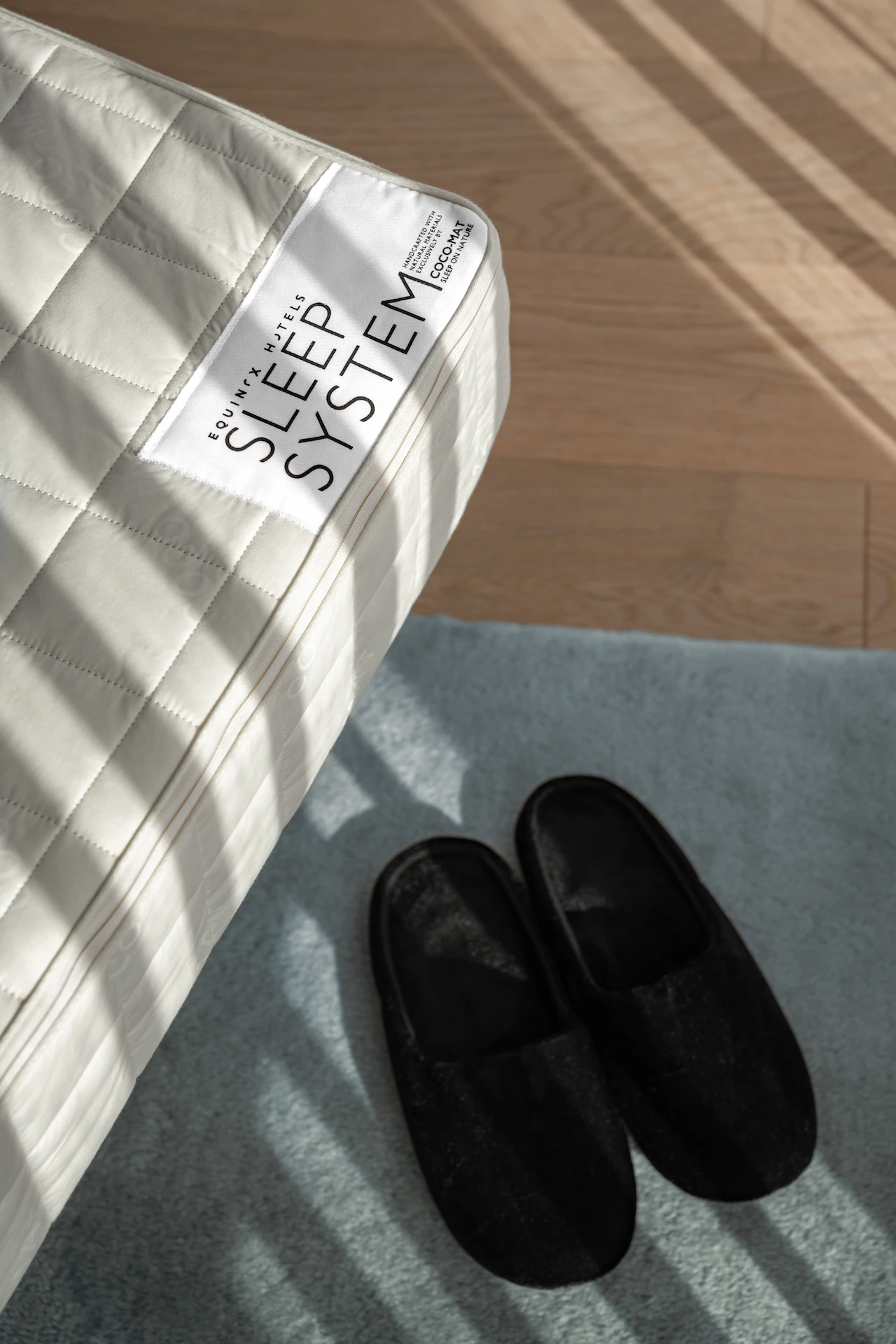 Equilibrium Series mattress with quilted surface and branded sleep system label, bathed in soft sunlight beside black slippers on blue rug
