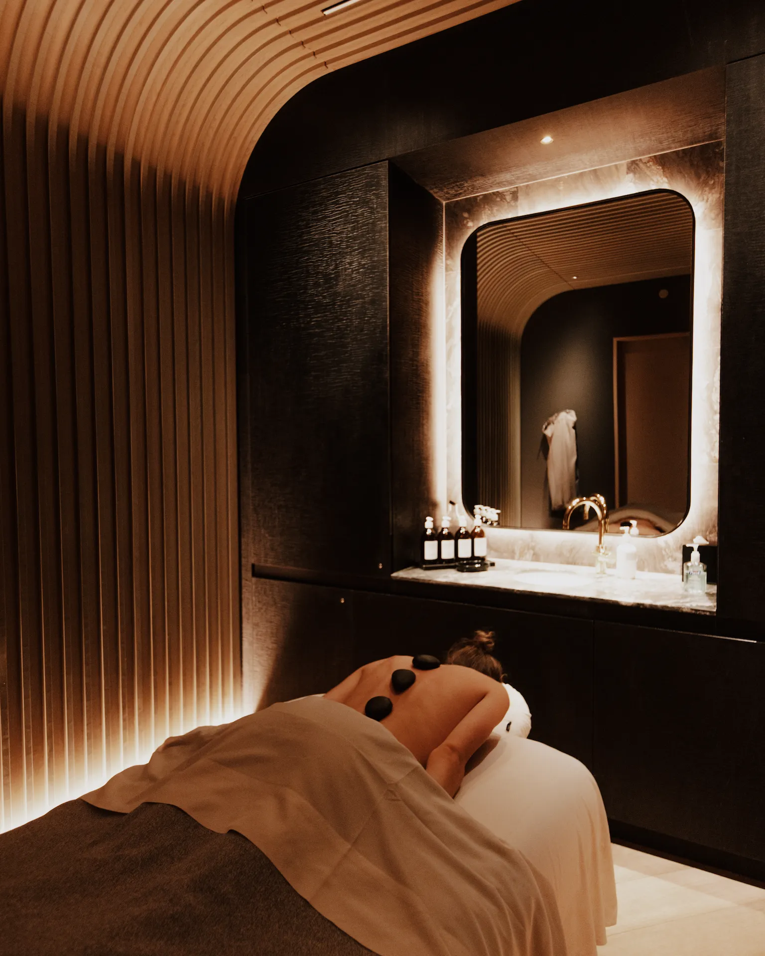 Guest receiving hot stone massage in serene spa room with curved wood paneling and marble accents