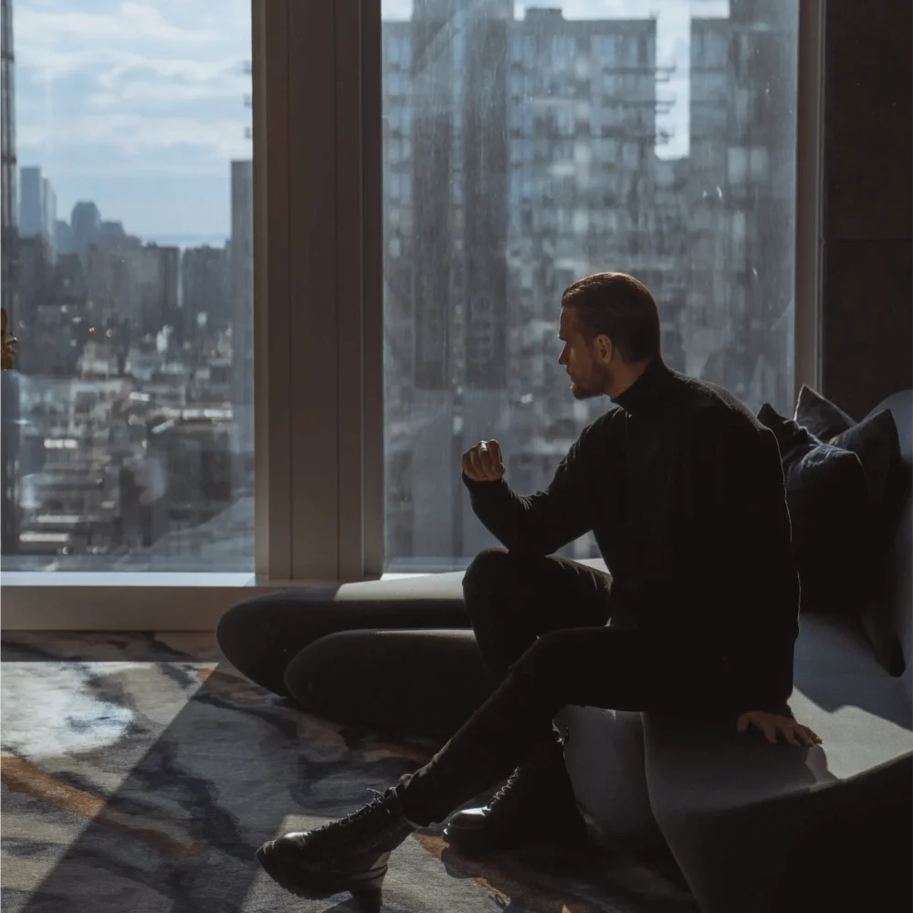 Person in dark clothing sitting on modern couch at Equinox Hotel, gazing out floor‑to‑ceiling windows at daytime cityscape.