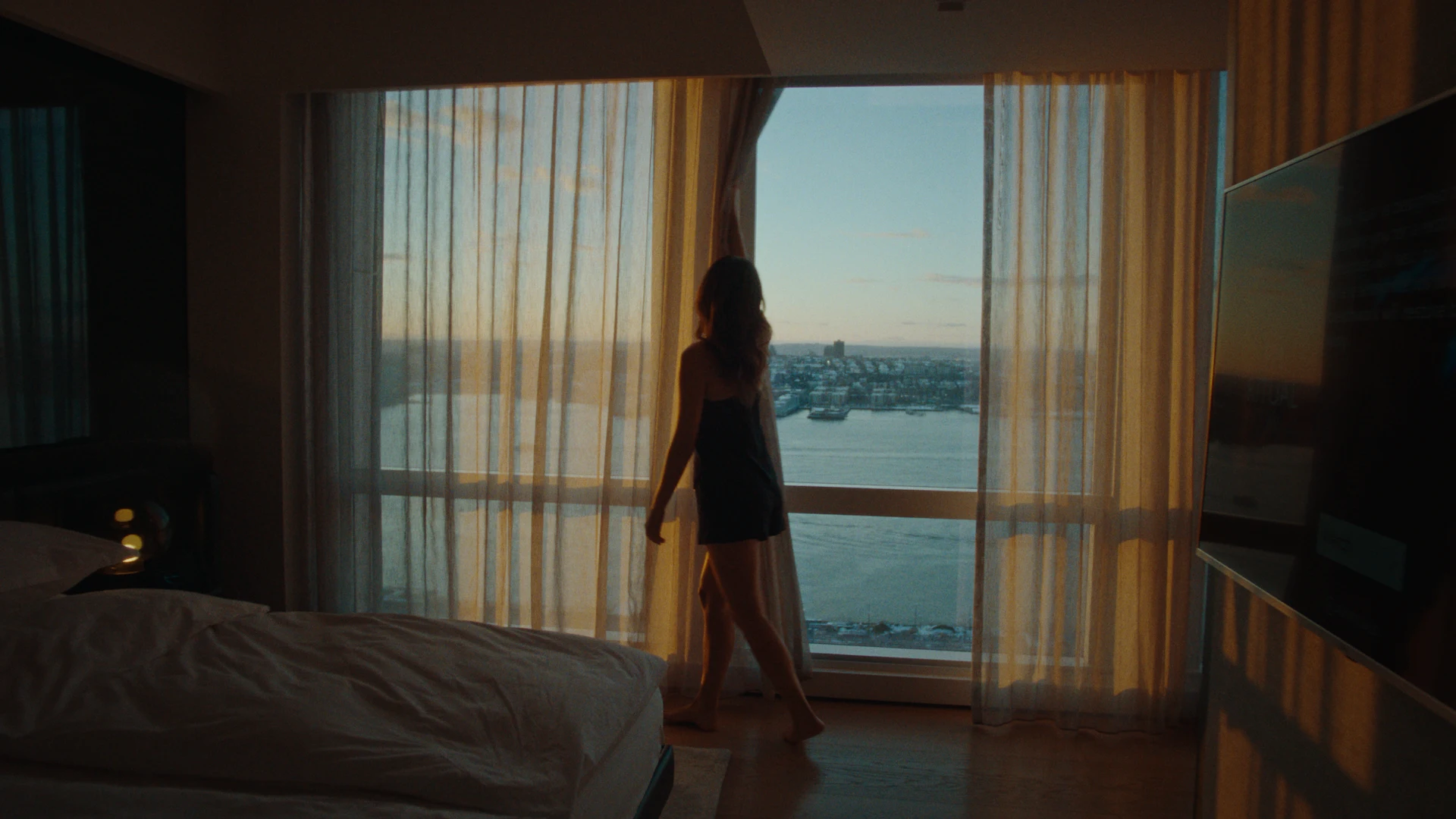 Hotel guest standing quietly before sheer-curtained window at sunrise, overlooking tranquil water and city skyline in a moment of reflection
