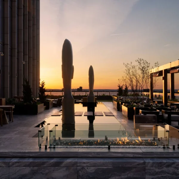 Rooftop terrace at sunset with fire pit, sculptures, and city views.