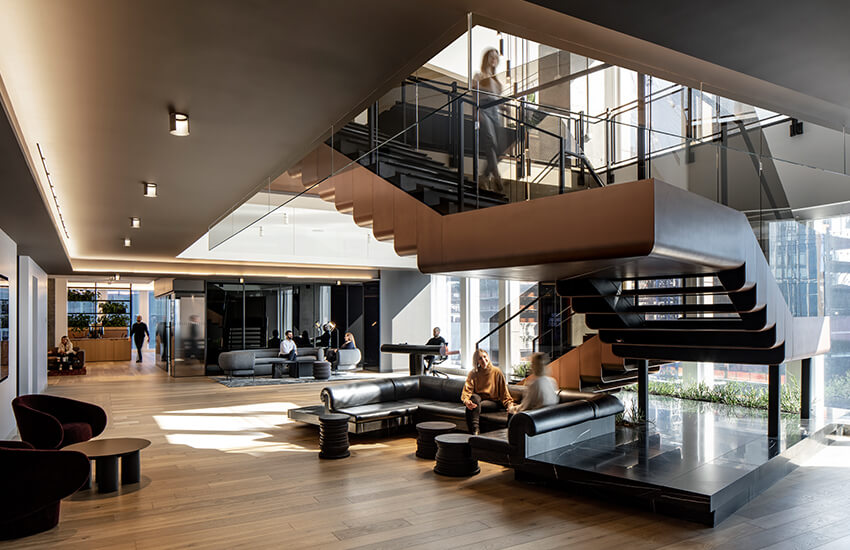 Modern office lobby with wood floors, glass staircase, large windows, and black seating areas.