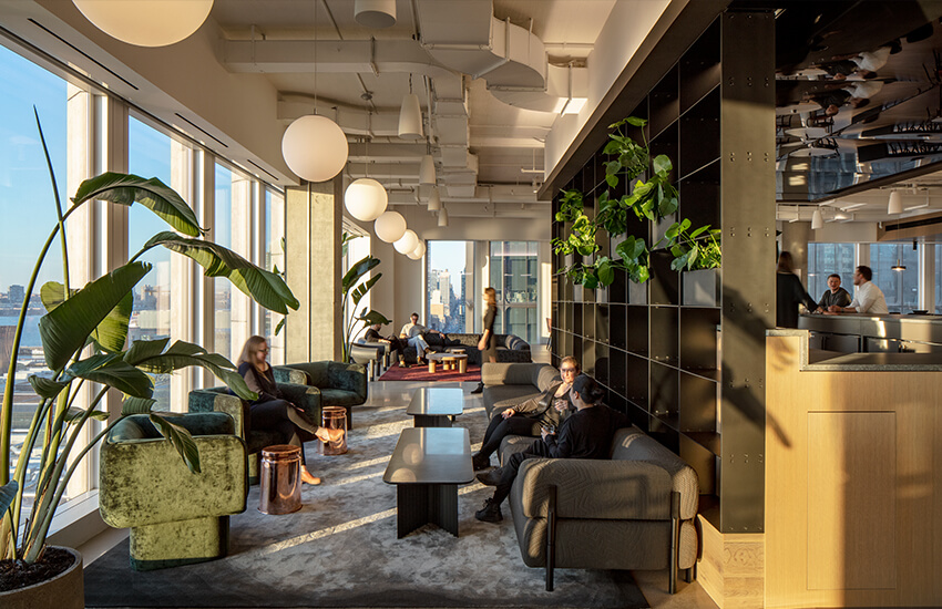 Sunlit lounge with large windows, modern seating, globe lights, plants, and people talking.