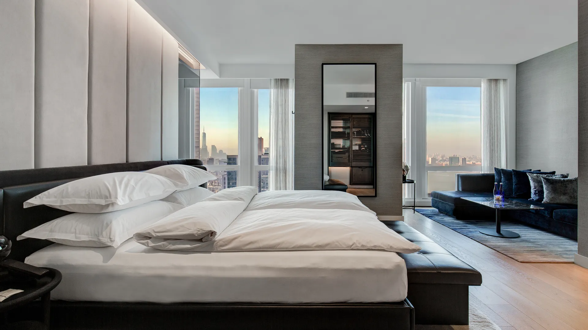 A guest room in charcoal and cream, a low bed against a padded leather headboard wall with a full-height mirror beside it and floor-to-ceiling windows onto the skyline.