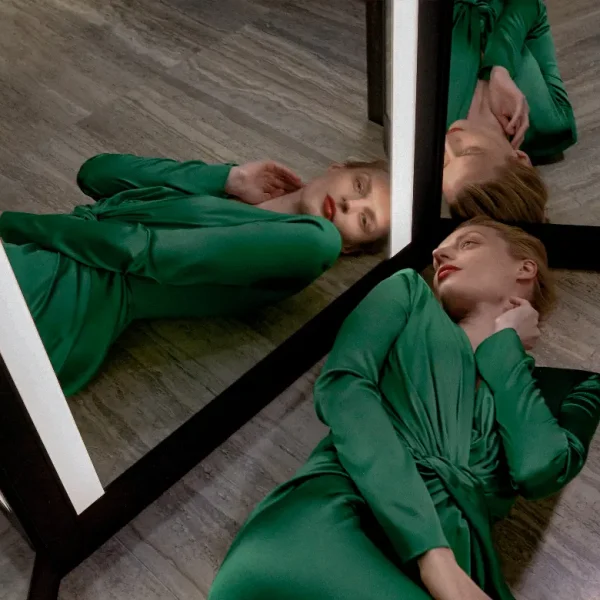 Woman in a green dress lying beside a large mirror, her reflection doubling the scene.