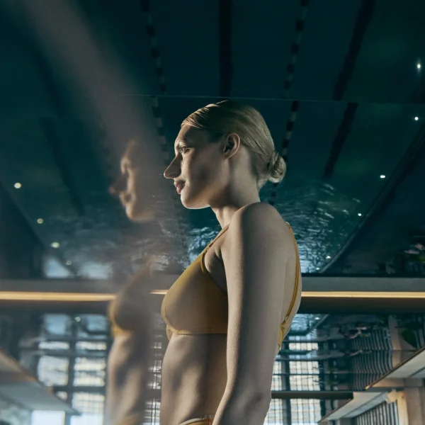 Woman in yellow bikini beside indoor pool, her reflection visible in sleek modern setting.
