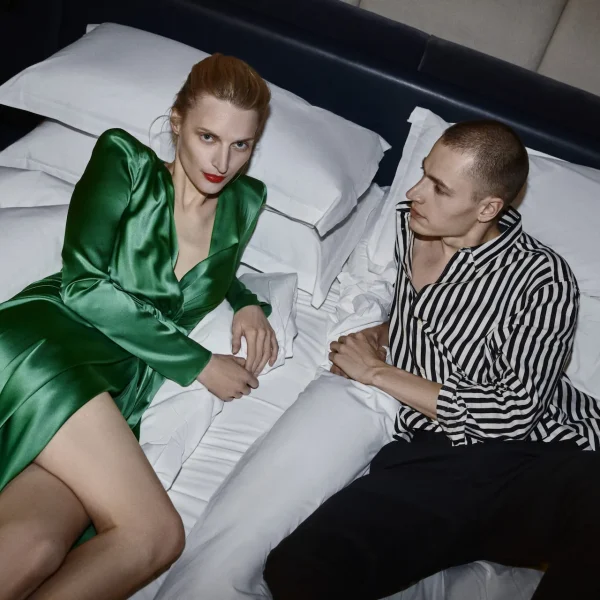 Two people lying on a bed facing each other, one in a green dress and one in a striped shirt.