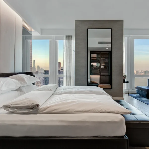 Modern luxury bedroom with large white bed, panel wall, sunset city windows, and seating area.