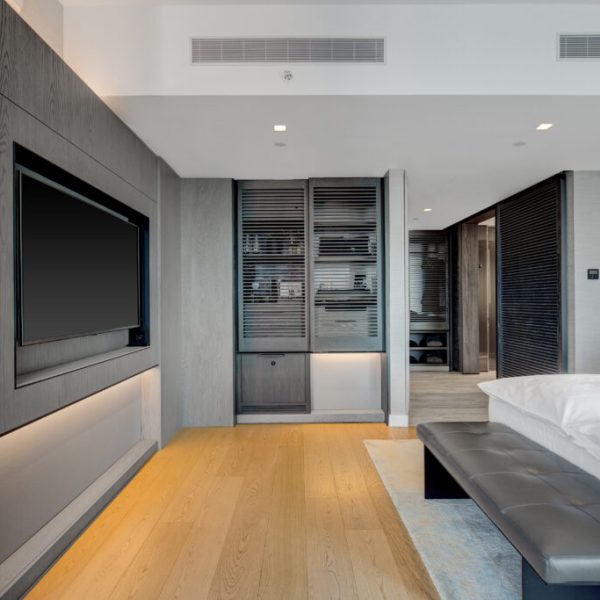 Minimal hotel bedroom with wall-mounted TV, white bed, padded bench, and soft hallway lighting.