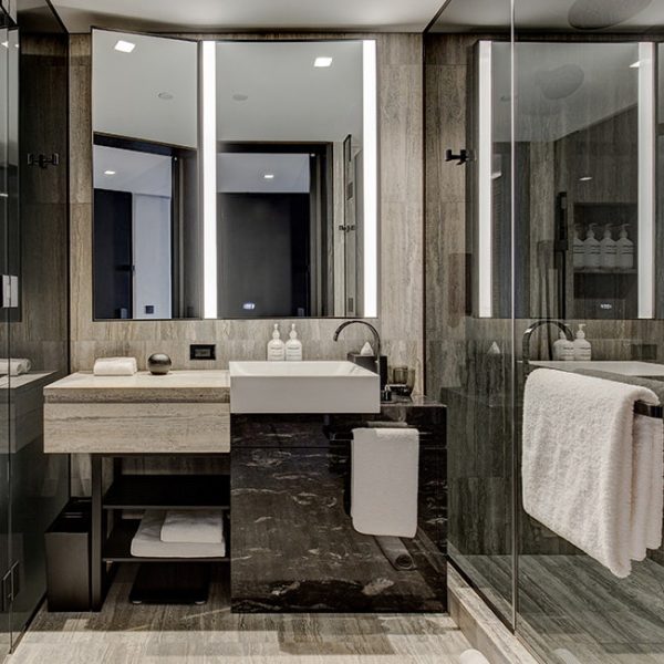A sleek, modern bathroom with neutral stone and black finishes, featuring a rectangular white vessel sink on a dark marble vanity. Tall backlit mirrors, a glass-enclosed shower with white towels, and neatly arranged amenities give the space a luxurious, hotel-like feel.