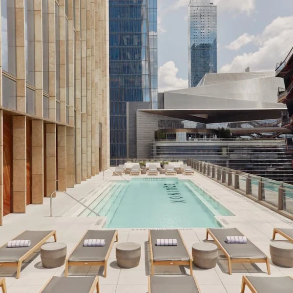 Sleek rooftop terrace with long rectangular pool, rows of lounge chairs, glass railings, and modern buildings.