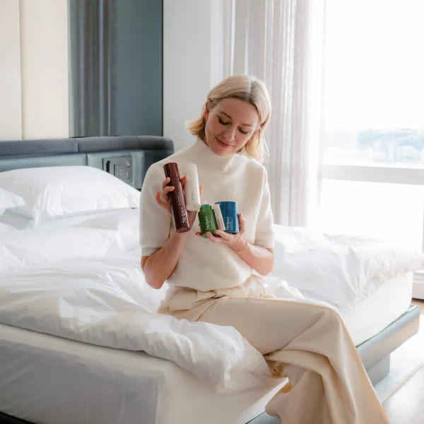 Woman in light clothing sits on white bed, smiling at colorful bottles in daylight.