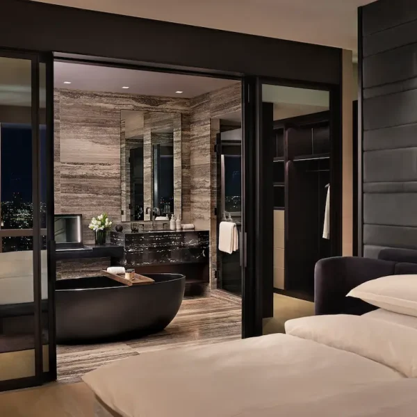 Luxurious hotel bedroom with glass doors revealing warm stone bathroom and sleek black bathtub.