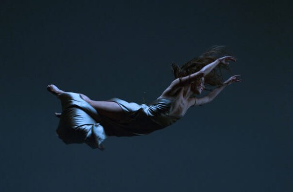 Person floating horizontally in darkness, wearing gown and holding pillow