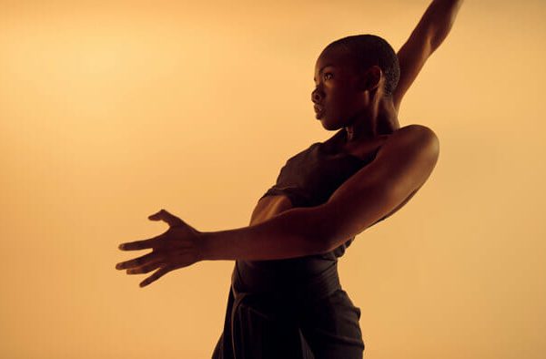Dancer in dark outfit extending arms mid-movement against warm golden background.
