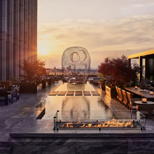Sunset rooftop terrace with long reflecting pool, wire-mesh head sculpture, lounge seating, and a fire line.