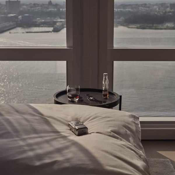 Unmade bed and bedside table with drinks, overlooking expansive river view through windows.