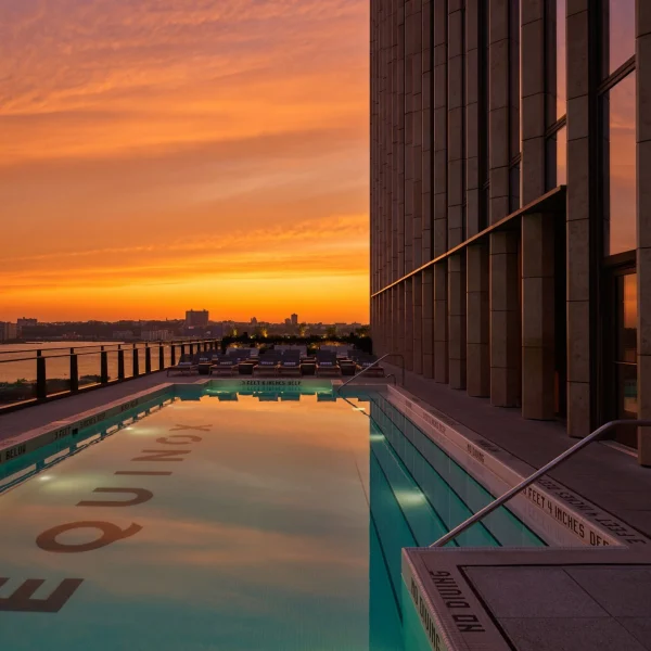 Rooftop pool beside a modern building reflects vivid sunset colors, lounges overlooking river skyline.