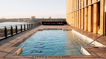Narrow rooftop pool with steps, glass railing, and wide river views in soft morning or evening light.