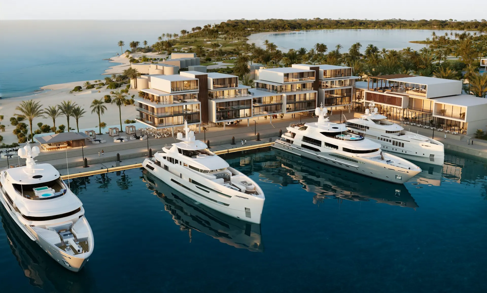 Modern waterfront development with glass-fronted buildings, docked yachts, sandy beach, and lush tropical landscaping.