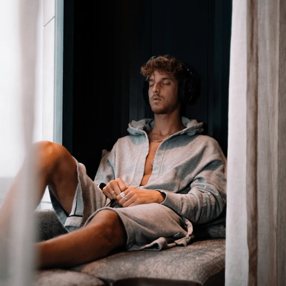Person in gray hoodie and shorts sitting by window with headphones, eyes closed in relaxed wellness moment