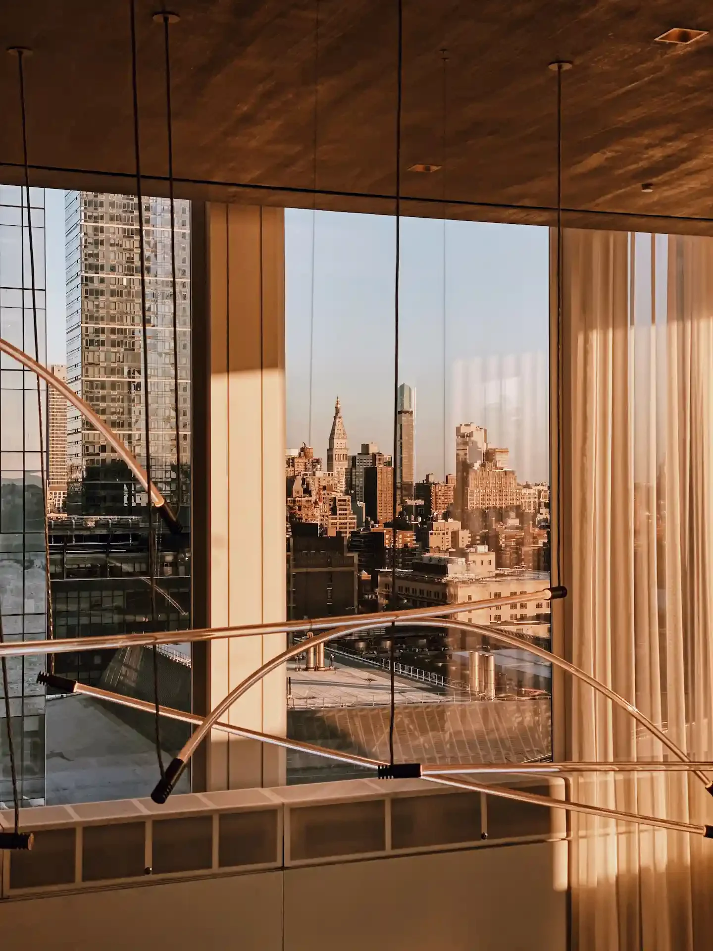 City skyline at golden hour seen through floor-to-ceiling windows with sheer curtains and sculptural light fixture