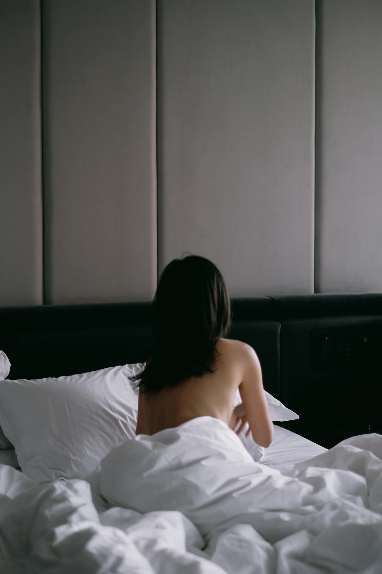 Person with long dark hair sitting on white bed, upper body unclothed, facing away from camera with neutral-toned backdrop.