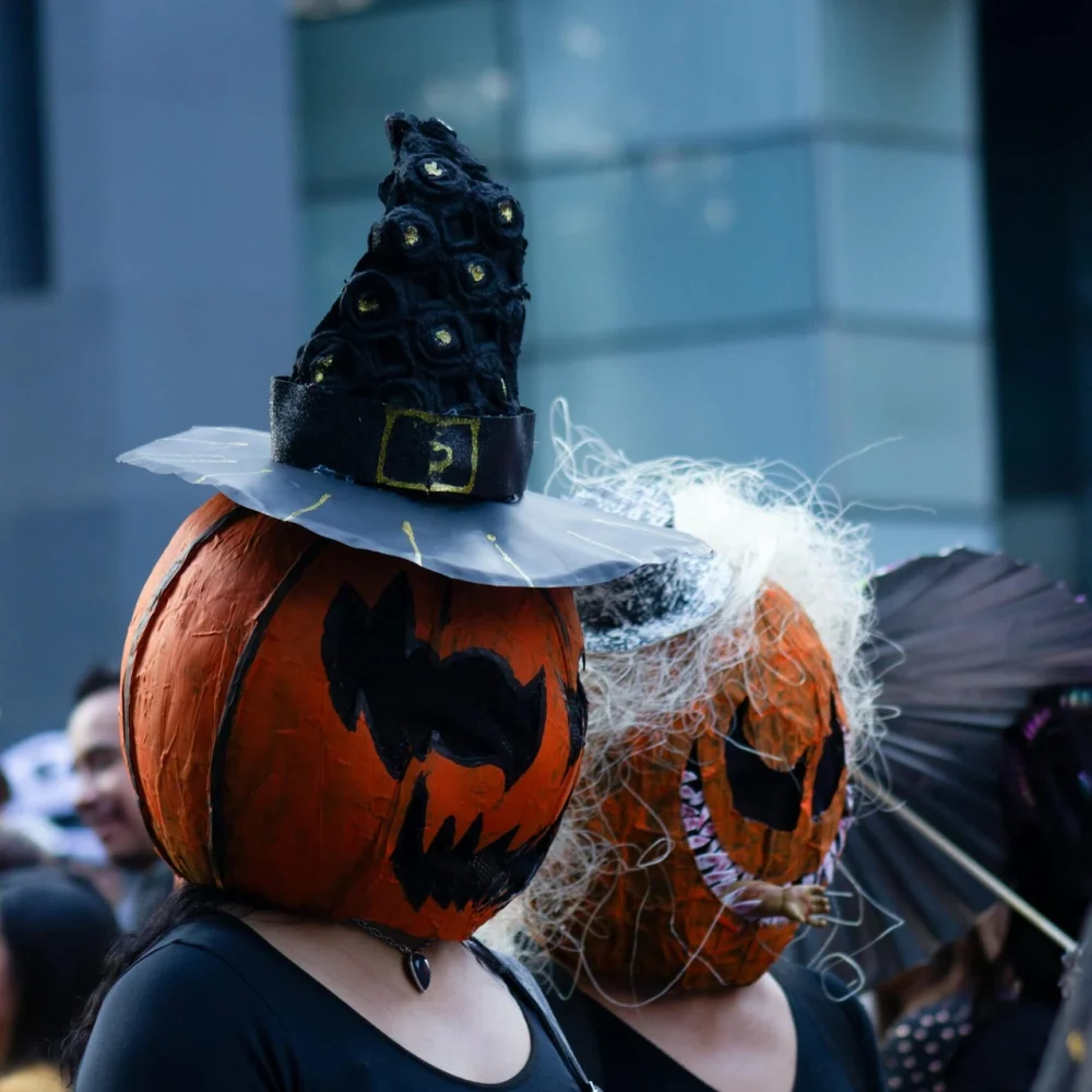 Two people in Halloween costumes with pumpkin heads, witch hat, curly wig, and black outfits in urban setting