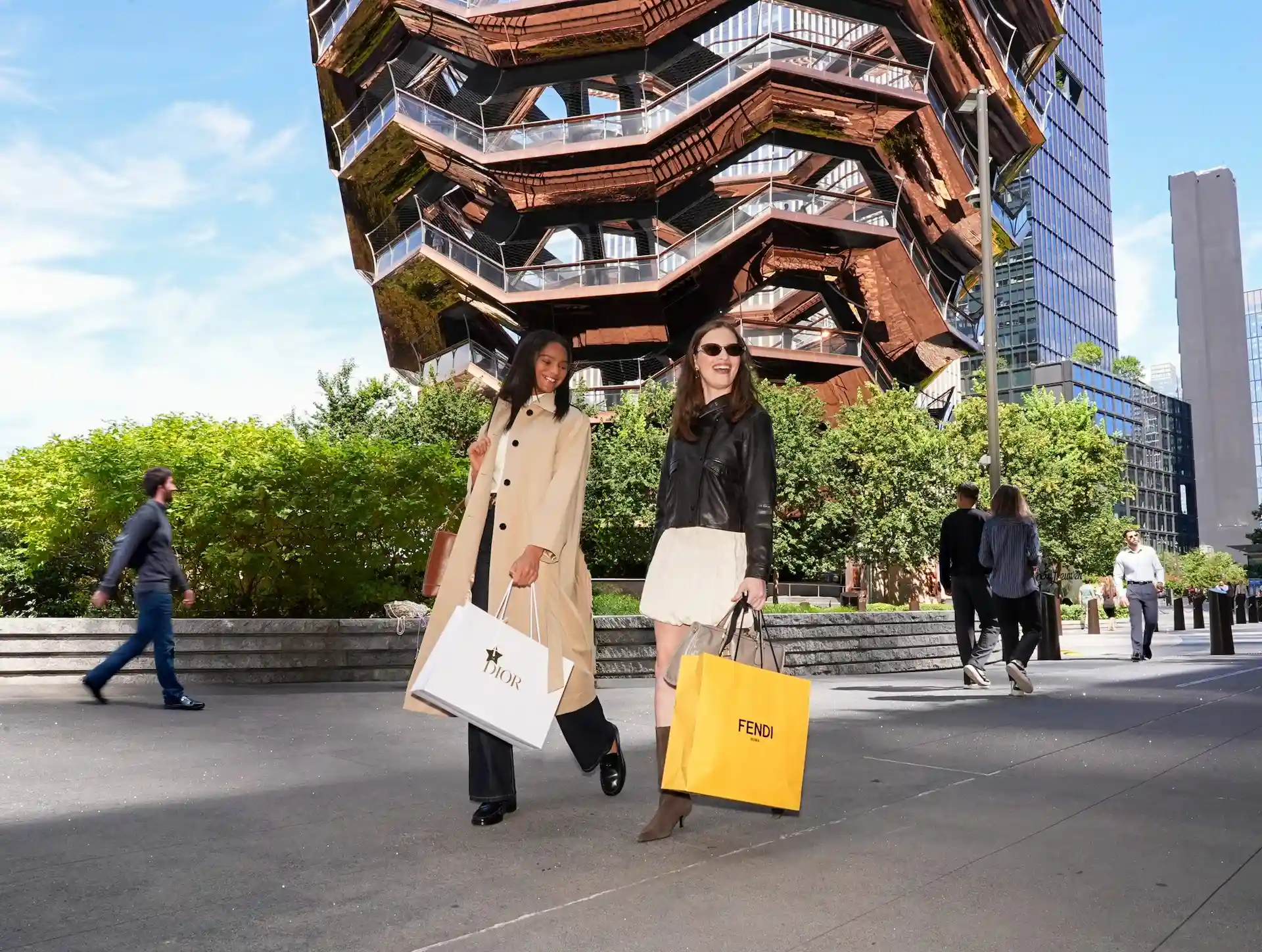 Two people with Dior and Fendi bags walking past the Vessel in Hudson Yards on a sunny day
