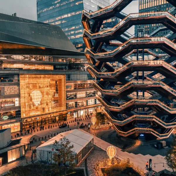 The Vessel at Hudson Yards lit with warm lights, surrounded by skyscrapers, people, and festive balloon displays.