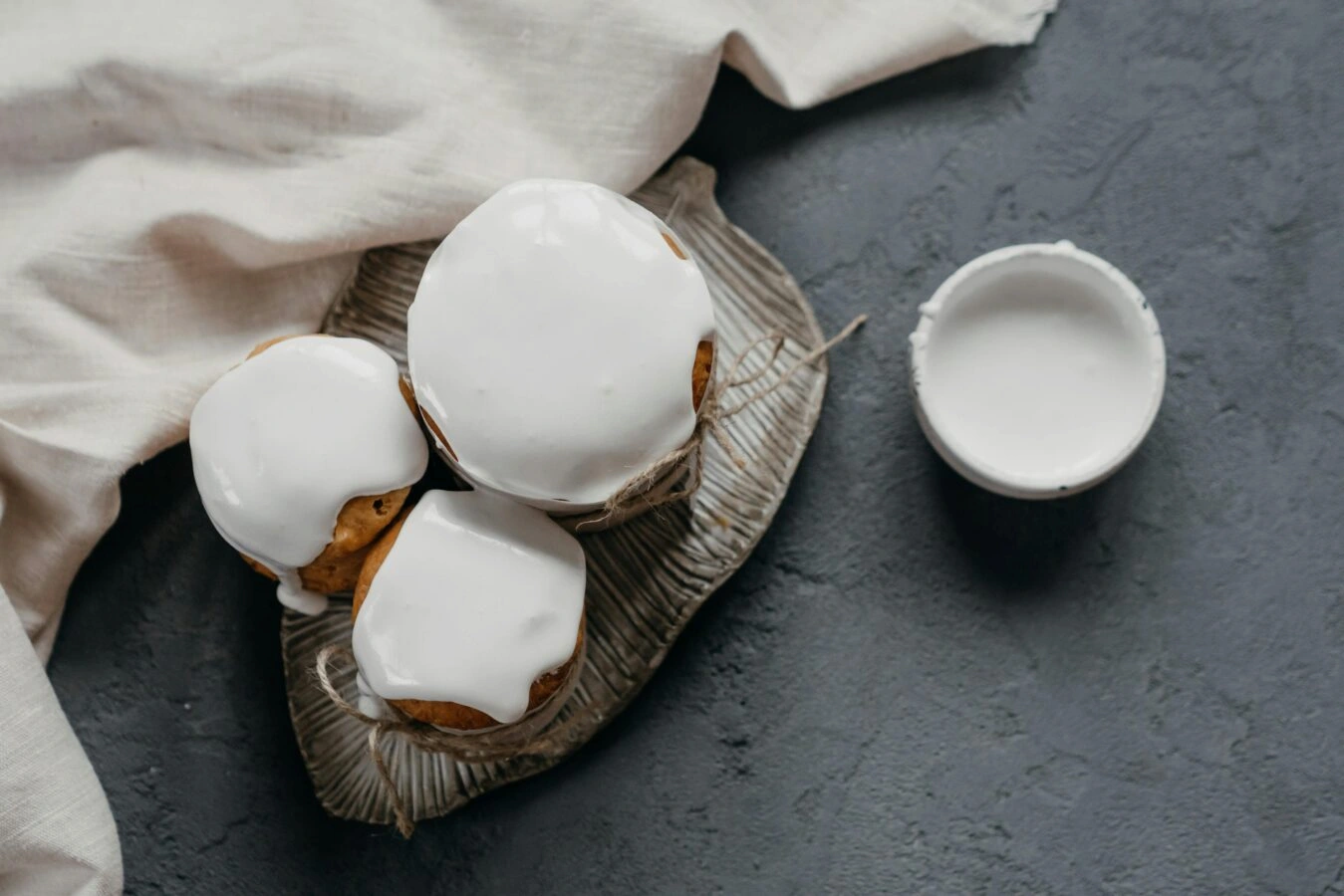 Three iced muffins on leaf-shaped plate with bowl of white frosting on dark textured surface
