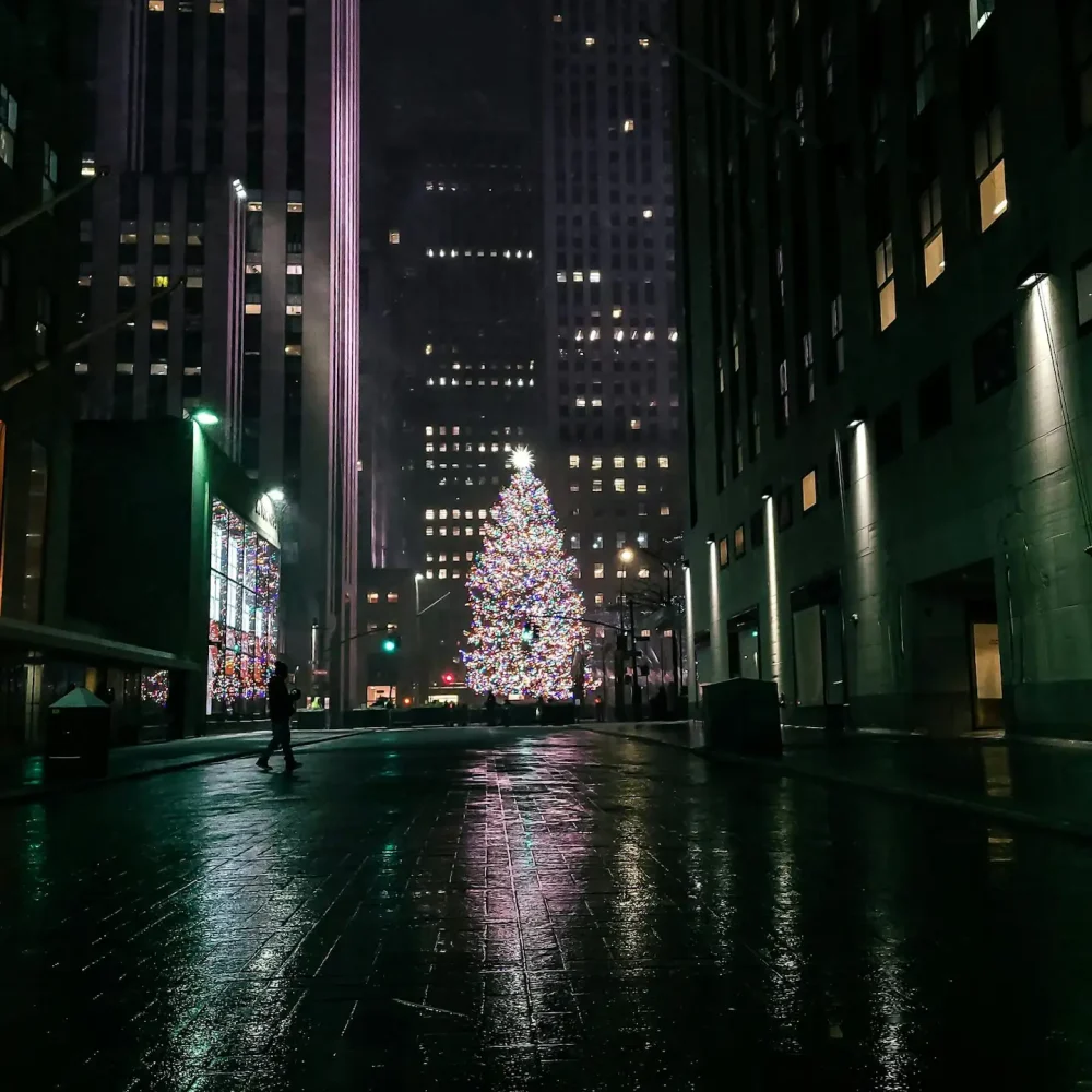 Large Christmas tree with multicolored lights and glowing star in city street at night with wet reflections